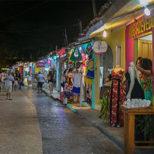 Calle peatonal empedrada con algunas personas caminando, rodeada de puestos de artesanías típicas como bolsos, ropa y frutas. Es de noche.