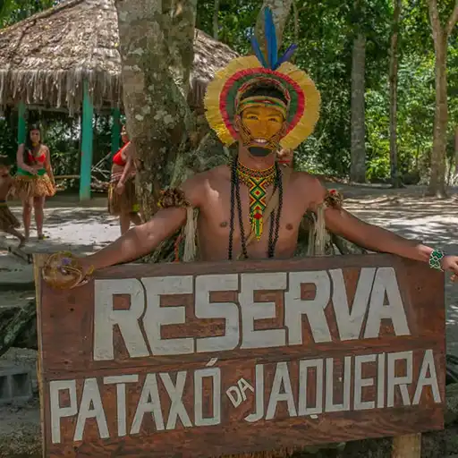 Indígena detrás de un cartel de madera que dice “Reserva Pataxó da Jaqueira” en la aldea Pataxó. Se observan otras personas de la comunidad y vegetación tupida.