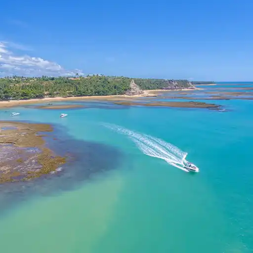 Vista panorámica de Praia do Espelho donde se observan embarcaciones, con una gran extensión de vegetación tupida de fondo. En primer plano, se observa el mar color turquesa. El día está soleado con algunas nubes.