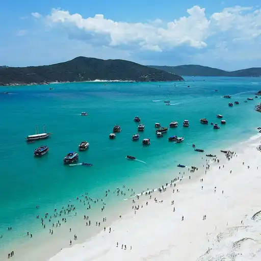 Vista panorámica de la playa en Arraial do Cabo, con personas disfrutando del día soleado, sobre la arena blanca y refrescándose en el mar turquesa, varias embarcaciones navegando y morros rodeando la playa.